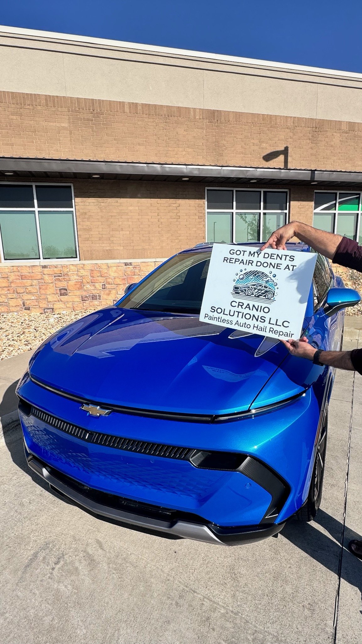 Customer holding a Cranio Hail sign over the hood of a repaired blue Chevrolet