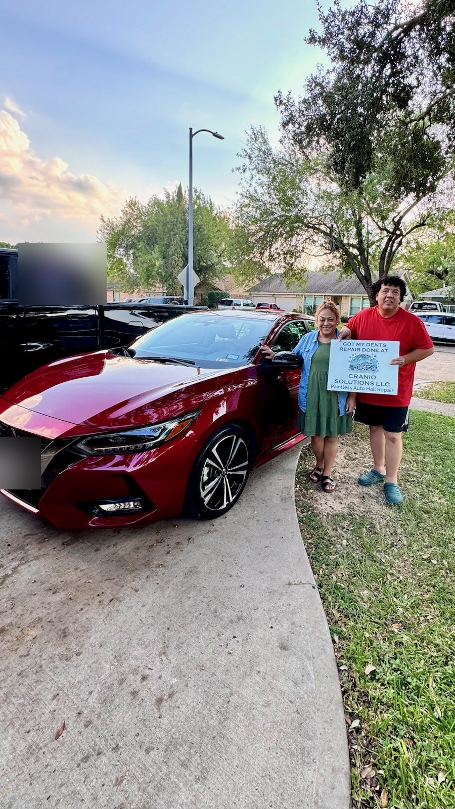 Two customers holding a Cranio Hail sign beside their repaired red Nissan